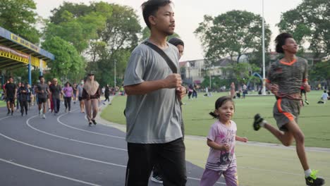 Menschen-Joggen-Auf-Einer-Strecke-In-Einem-Belebten-Park-Mit-üppigen-Bäumen-Und-Gebäuden-Im-Hintergrund