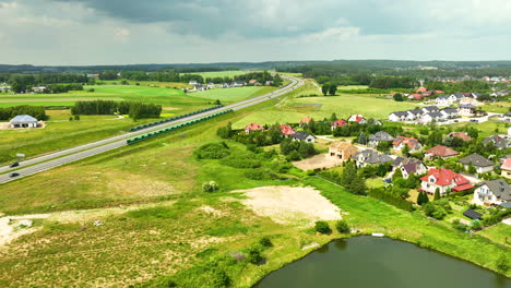 Aerial-view-of-a-highway-in-the-Polish-countryside,-featuring-nearby-residential-houses,-green-fields,-and-a-small-pond-under-a-partly-cloudy-sky