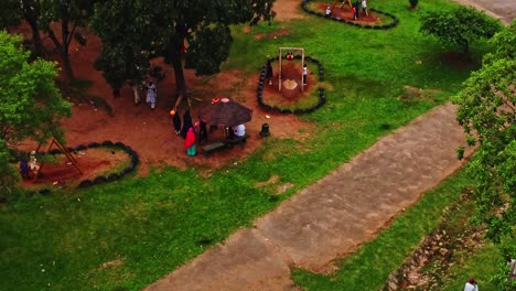 Jib-down-of-children-playing-on-swings-in-a-park-and-parents-roaming-through-the-green-grass-in-Abuja,-Nigeria