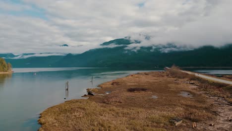 A-gravel-path-in-nature-leading-into-a-calm-peaceful-river-pathway-surrounded-by-water-yellow-brown-grass,-fog-mist-dark-green-mountains-clouds-in-fall-Aerial-approaching-Maple-Ridge-Pitt-Meadows-BC