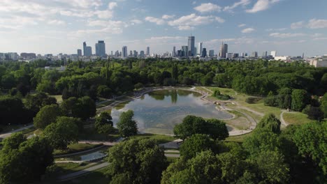 Impresionante-Horizonte-De-Varsovia-Sobre-Un-Parque-Verde-Con-Estanque-De-Agua,-Vista-Aérea