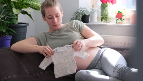 Pregnant-white-woman-sitting-on-the-couch,-playing-with-baby-clothes,-shirt-and-caressing-belly-slow-motion