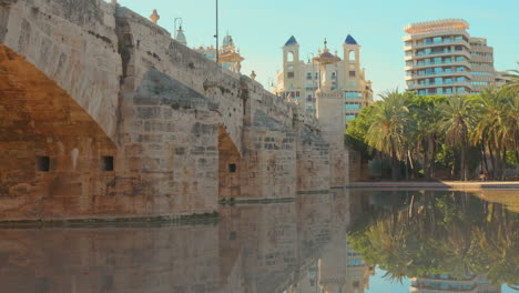 Historic-stone-bridge-over-the-serene-waters-in-Turia-Gardens-with-buildings-in-Valencia,-Spain