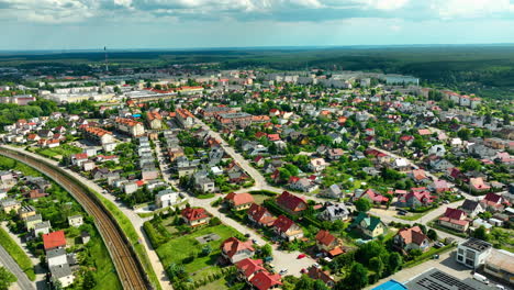 Aerial-view-of-Iława,-showcasing-a-sprawling-residential-area-with-a-mix-of-houses-and-apartment-buildings,-surrounded-by-greenery-and-intersected-by-a-railway-line-under-a-partly-cloudy-sky