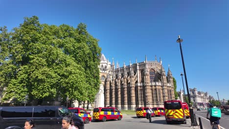 Westminster-Abbey-with-distinctive-architecture-located-on-Abingdon-Street,-flanked-by-red-police-vans-and-nestled-next-to-a-large-green-tree-on-a-sunny-day