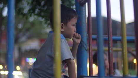 A-boy-playing-on-colorful-metal-bars-in-a-park-at-dusk,-looking-thoughtful-and-pensive