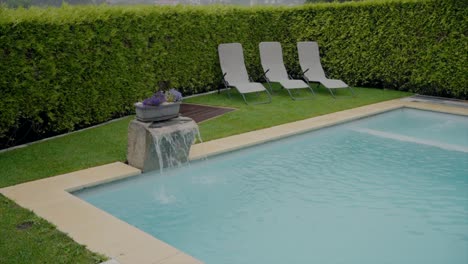 Outdoor-swimming-pool-with-three-lounge-chairs-and-a-small-water-feature-in-a-manicured-garden