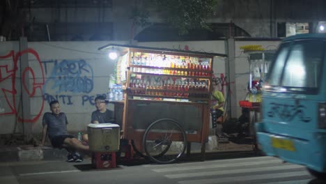 Two-men-sitting-by-a-lit-up-street-food-cart-at-night-in-Alun-Alun-Ahmad-Yani,-Tangerang