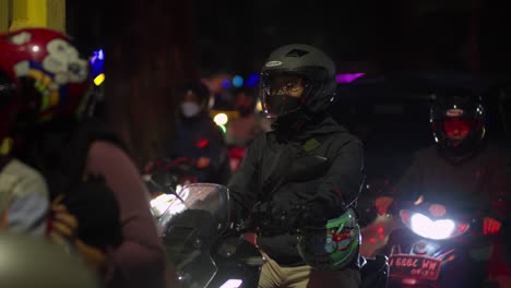 Motorcycle-riders-with-helmets-waiting-at-night-in-a-crowded-street,-illuminated-by-headlights