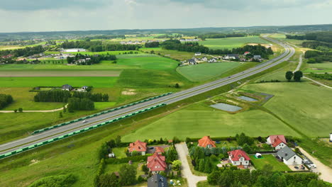 Aerial-view-of-a-highway-in-the-Polish-countryside,-surrounded-by-houses-and-green-fields-under-a-partly-cloudy-sky