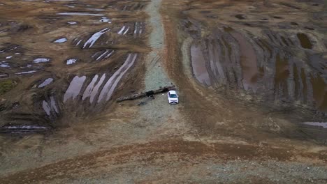 Drone-making-a-wide-orbit-around-a-white-car-parked-on-a-dark-muddy-flat,-with-intense-sky-reflections-in-puddles-and-off-road-vehicle-tracks