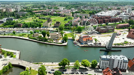 Aerial-view-of-Gdańsk,-showcasing-a-vibrant-cityscape-with-a-ferris-wheel,-river,-and-urban-greenery,-blending-modern-architecture-and-historic-charm