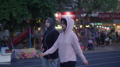 Dos-Mujeres-Con-Hijabs-Caminan-Juntas-Al-Atardecer-En-Un-Parque-Concurrido-Con-Gente-Y-Luces-Alrededor