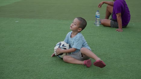 Young-boy-playing-with-a-soccer-ball-on-a-green-field,-supervised-by-an-adult