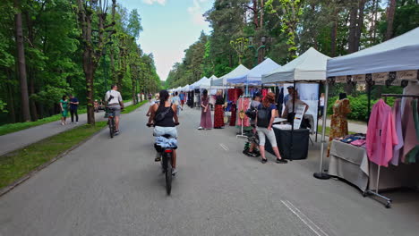 Slow-motion-shot-of-cyclists-riding-through-a-lively-outdoor-market-event-in-Mežaparks-in-Riga,-Latvia-on-a-cloudy-day