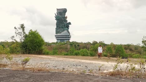 A-woman-with-her-dog-strolls-through-the-grounds-of-the-Garuda-Wisnu-Kencana-Cultural-Park,-Bali-on-a-cloudy-day