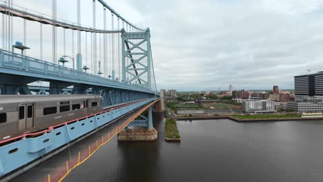 Toma-Aérea-De-Seguimiento-Del-Metro-De-Tren-Americano-En-El-Puente-Ben-Franklin-Durante-Un-Día-Nublado-En-Filadelfia