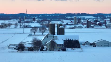 Aufsteigende-Drohnenaufnahme-Einer-Milchfarm-Im-Winterschnee-Bei-Sonnenuntergang