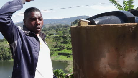 Hombre-Africano-Explicando-Cómo-La-Aldea-Recolecta-Agua-De-Lluvia-Con-Un-Tanque-De-Agua-En-Uganda,-África