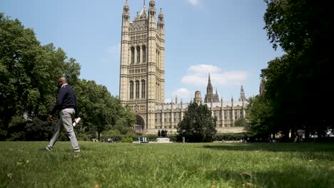 Se-Ve-A-Una-Persona-Caminando-Sobre-Un-Césped-Verde-Cerca-De-Las-Emblemáticas-Casas-Del-Parlamento-De-Londres-En-Un-Día-Soleado,-Encapsulando-La-Yuxtaposición-De-Naturaleza-E-Historia.