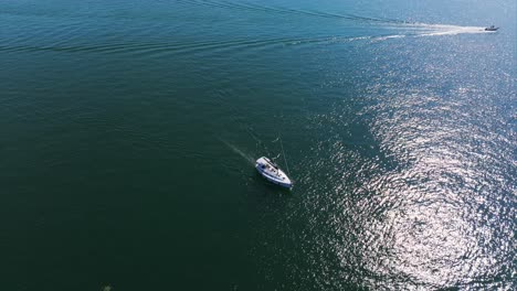 Aerial-overhead-view-of-lone-sailboat-cruises-across-the-expansive,-calm-waters-of-the-sea-under-a-clear-blue-sky,-leaving-gentle-waves-in-its-wake