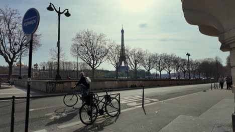 Toma-En-ángulo-Fijo-De-Un-Ciclista-Que-Pasa-Por-La-Carretera-De-París-Y-La-Famosa-Torre-Eiffel-Al-Fondo-En-Un-Día-Soleado---París,-Francia