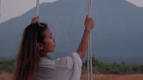 Woman-swings-in-Bali-savanna-with-mountain-backdrop-during-sunset,-reflecting-a-serene-moment