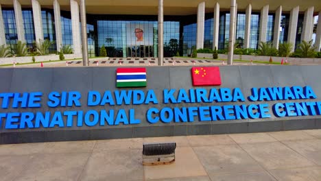 Banderas-Y-Letras-Del-Centro-De-Conferencias-Sir-Dk-Jawara-En-La-Entrada-En-Gambia,-África,-Vista-Panorámica-De-Izquierda-A-Derecha-En-Cámara-Lenta