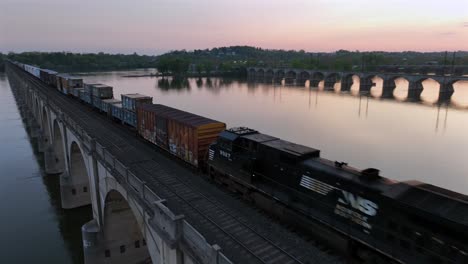 FPV-drone-flight-over-industrial-cargo-train-on-bridge-crossing-river-at-golden-sunset