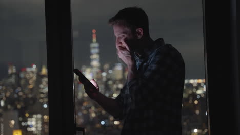 Un-Hombre-O-Un-Hombre-De-Negocios-Sosteniendo-Un-Teléfono-Inteligente-Leyendo-Y-Mirando-La-Pantalla-Por-La-Noche-Con-Los-Edificios-De-La-Ciudad-Metropolitana-Al-Fondo