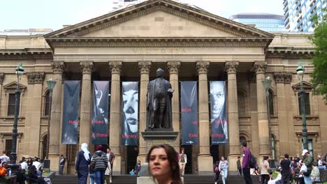 Die-Große-Fassade-Der-State-Library-Victoria-Mit-Der-Statue-Von-Sir-Redmond-Barry-Auf-Dem-Vorplatz,-Auf-Der-Sich-Einheimische-Und-Besucher-Auf-Der-Rasenfläche-In-Der-Stadt-Melbourne-Aufhalten