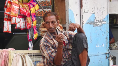 Middle-aged-man-sitting-on-a-bench-outside-his-small-shop-plucks-his-teeth-and-looks-in-the-camera-cautiously