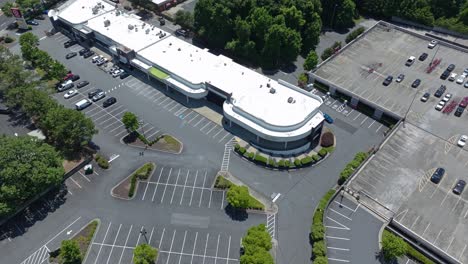 Inclinación-Aérea-Hacia-Abajo-Del-Centro-Comercial-Con-Estacionamiento-De-Autos-En-La-Ciudad-Americana-Durante-El-Día-Soleado