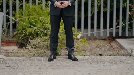 Man-in-a-black-suit-walking-confidently-on-a-path-with-greenery-and-a-fence-in-the-background