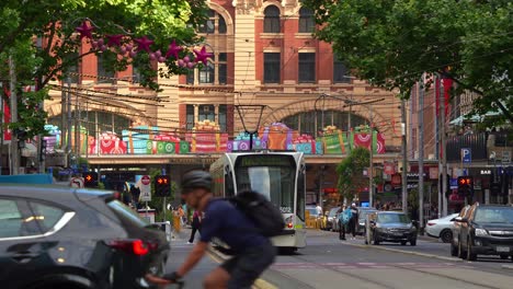 Vehicle-traffic,-pedestrians-crossing-and-trams-running-on-Elizabeth-street-with-tree-lined-street-decorated-with-festive-decorations-in-the-bustling-city-of-Melbourne,-a-vibrant-urban-scene