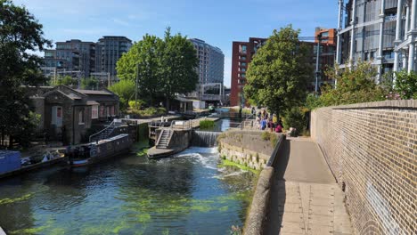 A-houseboat-entering-St-Pancras-Lock-on-Regent's-Canal-with-surrounding-urban-landscape-and-buildings,-King's-Cross,-London,-UK,-July-2023