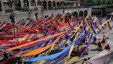 Colorful-ribbons-displayed-over-the-courtyard-of-Coal-Drops-Yard-with-people-relaxing-underneath,-King's-Cross,-London,-UK,-July-2023