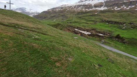 Aerial-FPV-view-of-a-hiker-walking-on-a-green-mountain-meadow-with-a-wooden-cross-and-a-small-church-in-the-background