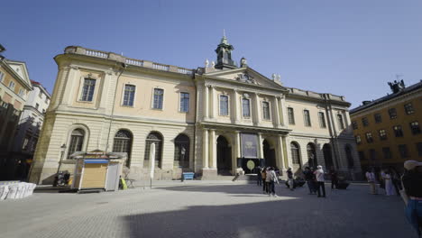 Exterior-of-the-Nobel-Prize-museum-at-midday-with-tourists-exploring-city,-pan-right