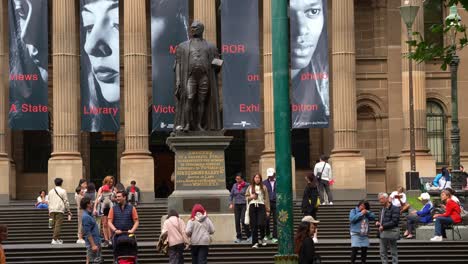 The-State-Library-Victoria-with-Corinthian-columns,-featuring-the-statue-of-Sir-Redmond-Barry-located-on-the-forecourt,-tourists-visiting-the-landmark-building,-locals-hanging-out-on-the-lawn