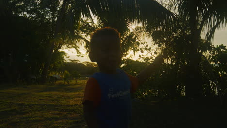 Young-Fijian-child-backlit-against-silhouette-of-palm-trees-raises-hand-point-out-to-right