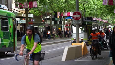 Enjambres-De-Ciclistas-Y-Scooters-Viajan-En-Su-Carril-Exclusivo,-Junto-A-Las-Vías-Del-Tranvía-Por-Donde-Circulan-Los-Tranvías,-En-La-Ciudad-De-Melbourne,-Destacando-El-Entorno-Urbano-Dinámico-Y-Verde-De-La-Ciudad.