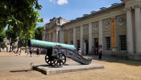 Un-Cañón-Histórico-Exhibido-Fuera-Del-Centro-De-Visitantes-Del-Antiguo-Royal-Naval-College-Con-Visitantes-Cercanos,-Greenwich,-Londres,-Reino-Unido,-Julio-De-2023