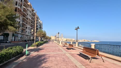 Walking-along-the-path-near-Malta's-coastline,-with-views-of-coastal-buildings