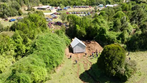 Aerial-View-Of-Excavator-pulling-small-house-nestled-in-a-lush-green-valley-surrounded-by-forested-hills-In-Chiloe,-Chile