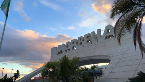 Big-Marbella-sign-with-flags-and-sunset-clouds-in-Spain,-orange-sky-and-palm-trees,-4K-shot