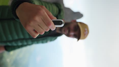 Hiker-wearing-a-green-jacket-and-a-brown-hat-is-holding-a-small-action-camera,-while-standing-in-a-scenic,-mountainous-area-waving-a-hand