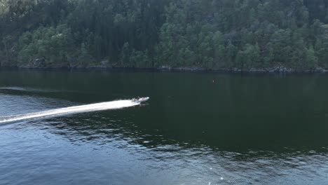 Drone-captures-rib-boat-cruising-from-behind-a-headland-in-a-Norway-fjord,-heading-towards-a-steep-mountain-wall