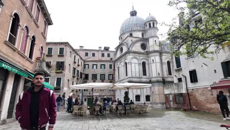 Strolling-across-a-small-square,-with-a-charming-coffee-shop-overlooking-the-grandeur-of-The-Church-of-Santa-Maria-dei-Miracoli-in-the-Cannaregio-sestiere-of-Venice,-Italy