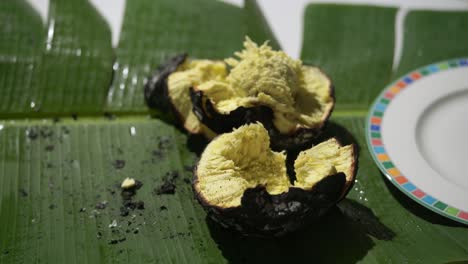 Roasted-breadfruit-in-open-fire,-man-spooning-out-the-cooked-roasted-breadfruit-on-banana-leaf-and-putting-in-plate,-traditional-Seychelles-food,-Mahe,-Seychelles-4k-24fps-008